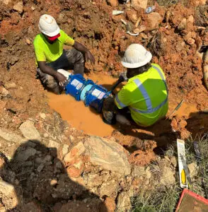 Two CWS utility workers in hard hats and safety gear repairing a water pipe in a muddy trench.