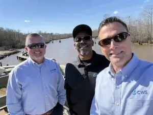 Three men in a selfie in front of a body of water and woods.