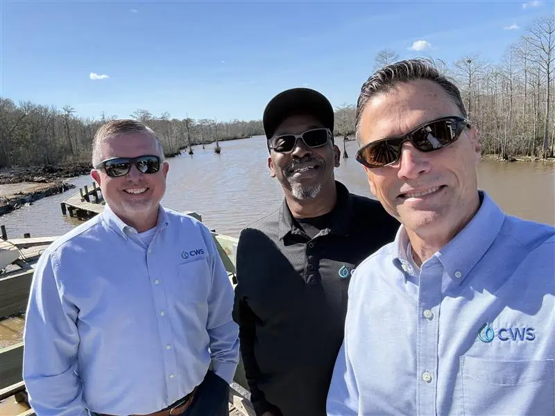 Three men in a selfie in front of a body of water and woods.