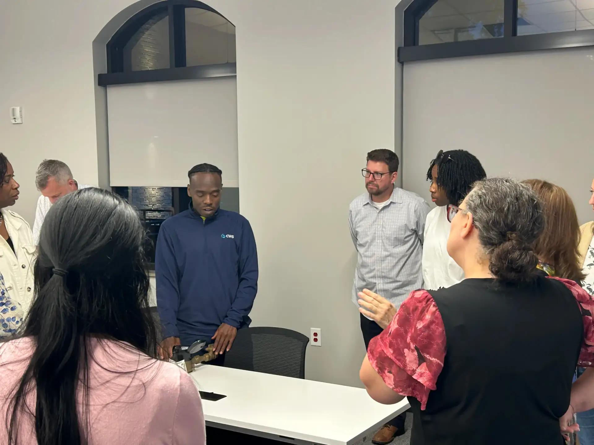 A group of people stand in a circle in a bright office room, watching a man in a navy CWS quarter-zip pullover demonstrate a brass water meter fitting on a white table.