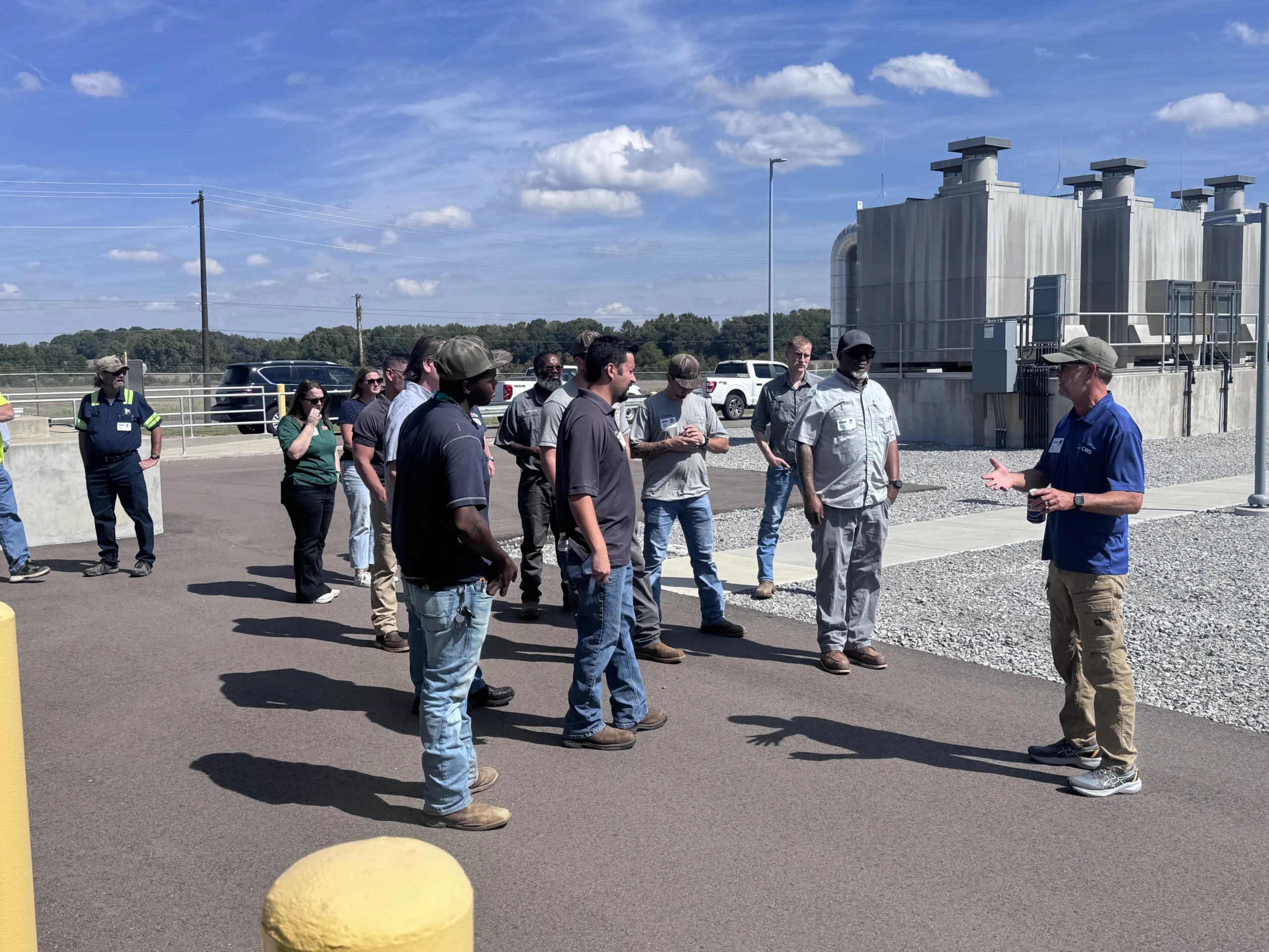 A man in a blue “CWS” polo gestures while speaking to a group of about a dozen people gathered outdoors on a paved lot beside a large concrete industrial facility.